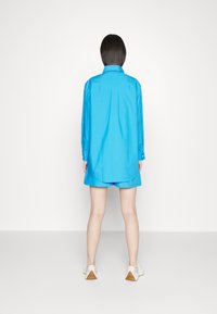 Blue long-sleeve shirt paired with matching shorts. The shirt features a collar, a back pleat, and buttoned cuffs. Sneakers with textured soles.