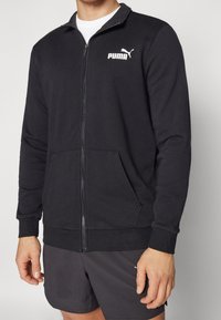 Giacca nera con zip, colletto alto, tasche frontali, polsini a costine e logo Puma bianco sul lato sinistro del petto. Tessuto morbido e strutturato.