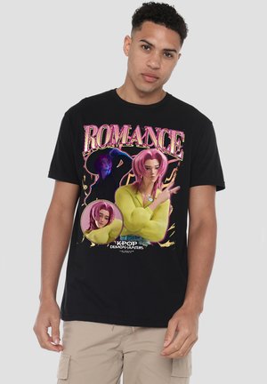 Young man wearing black T-shirt featuring "ROMANCE" text and animated characters with pink hair from Kpop Demon Hunters, beige pants.