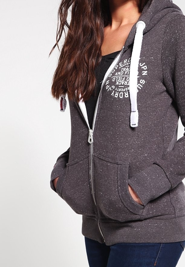 Sudadera gris con cremallera, hecha de una tela texturizada, con cordones blancos, bolsillos frontales y un estampado gráfico circular en el pecho.