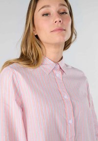 Light pink and white striped collared shirt with buttons, featuring long sleeves and a relaxed fit. Smooth fabric with a subtle sheen.