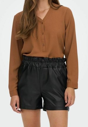 Kvinde iført en brun langærmet, knappet bluse, stoppet ned i højtaljede sorte shorts i kunstlæder med flæsekant ved linningen.