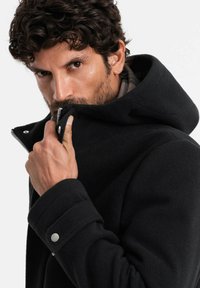 Abrigo de lana negro con capucha, que incluye un cierre con cremallera, botones a presión y un acabado texturizado. Detalles destacables incluyen un cuello en ángulo y puños.