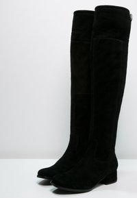 Botas negras sobre la rodilla de ante, con un diseño sólido, puntera redondeada y un tacón grueso para mayor estabilidad. Textura suave.