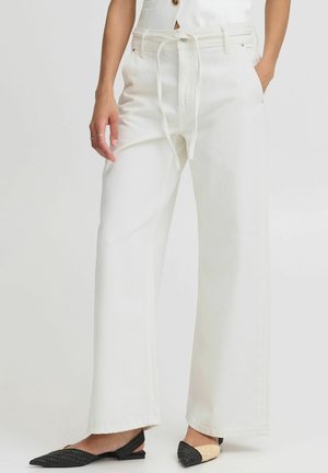 Kvinde iført hvide vidde bukser med højtaljet stofbælte, kombineret med sorte og beige vævede slingback-flats.