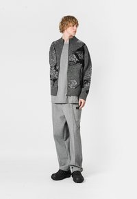 Cardigan gris de punto con diseño floral, camiseta gris, pantalones deportivos grises y calzado negro. Materiales con textura, corte relajado, detalle visible de bolsillo.