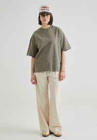 Tricou scurt cu mânecă scurtă de culoare verde măsliniu, cu o croială relaxată, asortat cu pantaloni largi de culoare crem și adidași bej. Șapca prezintă text roșu pe un fundal deschis.