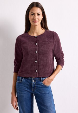 Kvinde med langt mørkt hår iført en knappet lilla kordfløjlsjakke og blå jeans med den ene hånd i lommen, stående mod en lys baggrund.