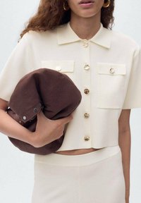 Blusa de manga corta color crema con botones dorados y dos bolsillos en el pecho, combinada con un bolso marrón texturizado, sostenido bajo el brazo de la persona.