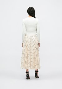 Wit, nauwsluitend topje met lange mouwen gecombineerd met een beige geplooide rok met zwarte stippen. Model draagt zwarte enkellaarzen met riempje.