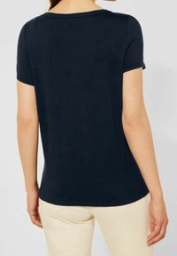 Granatowa koszulka polo z krótkim rękawem, okrągłym dekoltem, gładką fakturą i luźnym krojem, pokazana z tyłu, w połączeniu z jasnymi spodniami.