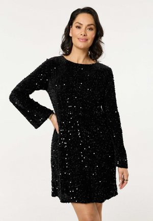 Woman wearing a long-sleeve black sequined dress standing with one hand on hip against plain background.