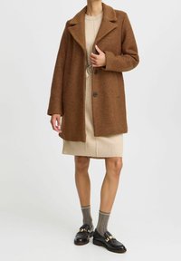 Bruin textuur jas met reverskraag, twee knopen, gedragen over een lichtbeige jurk. Zwarte loafers met gouden accenten en gem�êleerde sokken.