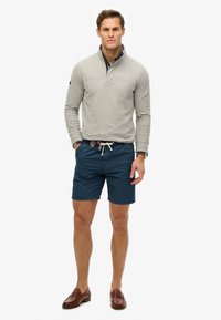 Pull gris à col zippé mi-hauteur, associé à un short bleu marine et des mocassins marron. Les détails notables incluent une taille avec cordon de serrage visible.