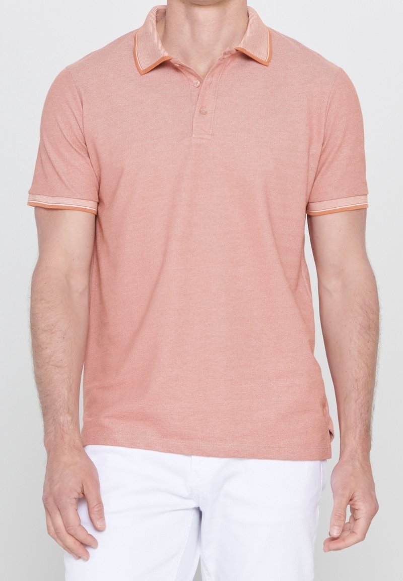 Homme portant un polo rose saumon à manches courtes et un pantalon blanc, debout devant un fond clair et uni.