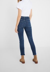 Mörkblå skinny jeans med hög midja, utrustade med bakfickor och lätt blekning. Matchas med beiga ankelskor mot en ljus bakgrund.