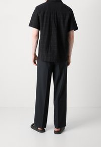Zwart shirt met korte mouwen en een gestructureerd ruitpatroon, gecombineerd met losvallende donkere broeken en zwarte sandalen.