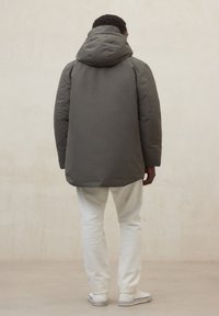 Giacca imbottita grigia con cappuccio, caratterizzata da una trama liscia e un design leggermente allungato. Indossata con pantaloni bianchi e scarpe casual.