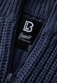 Jūras zila adīta džemperis ar biezu tekstūru, ar rāvējslēdzēju un markas etiķeti. Uz etiķetes redzams "Brandit Individual Wear".