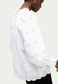 Blusa de algodón blanca con patrones bordados cortados, un cuello con volantes y puños elásticos con bordes festoneados.
