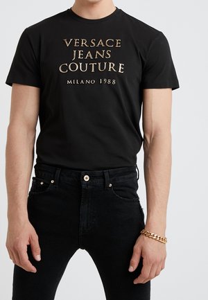 Mies, jolla on musta Versace Jeans Couture -t-paita kultaisella tekstillä ja mustat farkut, asusteena kultainen ketjurannekoru oikeassa ranteessa.