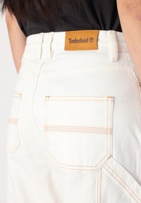 Bijele traperice od denima s narančastim šavovima, velikim stražnjim džepovima i smešnim kožnim Timberland logom na pojasu.