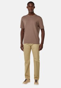 Camisa marrón de manga corta y pantalones beige. La camisa tiene un escote redondo; los pantalones tienen un corte recto. Unos zapatos de color claro completan el atuendo.