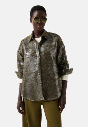 Persoon die een gouden paillettenshirt met knopen draagt, olijfgroene broek, wit shirt met lange mouwen eronder en ronde reflecterende zonnebrillen.