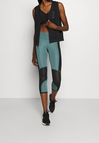 Musta hihaton liivi mustan urheilutopin päällä, yhdistettynä teal- ja mustiin capri-leggingseihin, joissa on geometrinen kuosi. Mustat lenkkarit viimeistelevät ilmeen.