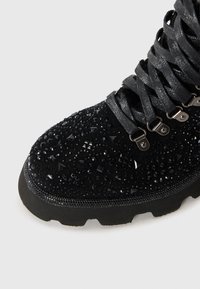 Stivale in suede nero con decorazioni testurizzate, accenti simili a cristalli e una suola in gomma robusta. I lacci sono grigi e intrecciati.
