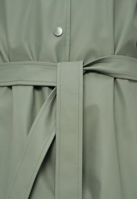 Κοντινή λήψη στη μέση ενός παλτό σε φαρδιάνη πράσινη απόχρωση, με ραμμένη ζώνη δεμένη μπροστά και ένα ματ μεταλλικό κουμπί πάνω από τη ζώνη.