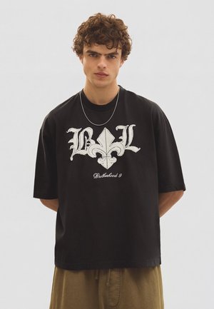 Jonge man met krullend haar die een zwart oversized T-shirt draagt met witte decoratieve tekst en embleem, een bruine broek en een dunne ketting.