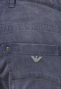 Granatowe spodnie dżinsowe z tylną kieszenią z logo srebrnego orła oraz metalowym zapięciem. Widoczne przeszycia wzdłuż krawędzi kieszeni.