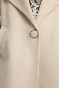 Cappotto di lana beige con ampio colletto e un grande bottone rotondo dorato, che mostra parte di un indumento con motivo sottostante.