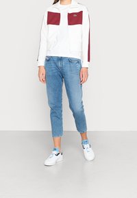 En person klädd i en vit och vinröd zip-up jacka, vit t-shirt, ljusblå cropped jeans och vita sneakers med blå detaljer.