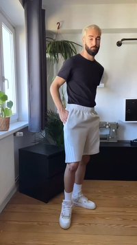 Man with light hair and beard stands indoors wearing black shirt, white shorts, white socks, and white sneakers near plants and sewing machine.