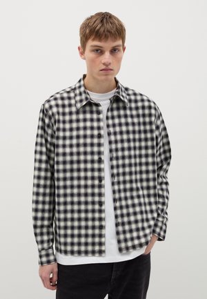 Chemise à carreaux noire et blanche à boutons, avec une coupe décontractée, un col et des manches longues, portée par-dessus un t-shirt blanc.