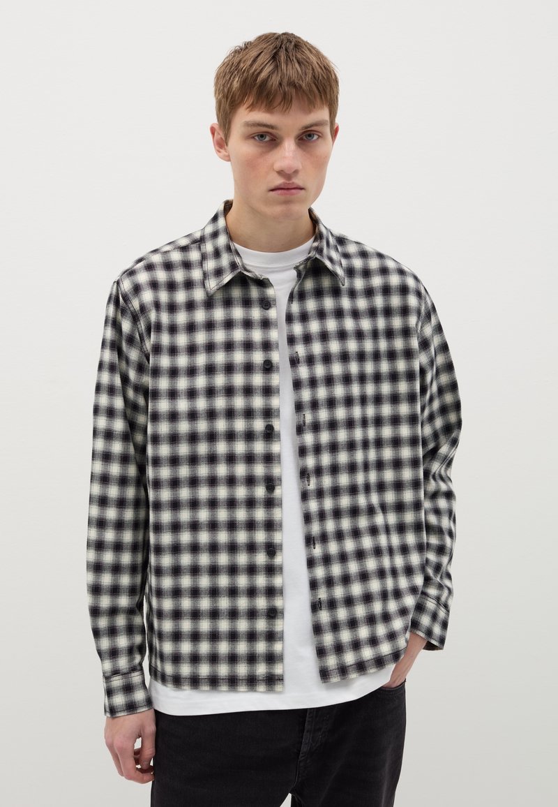 Chemise à carreaux noire et blanche à boutons, avec une coupe décontractée, un col et des manches longues, portée par-dessus un t-shirt blanc.