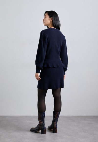 Vestido de punto azul marino con cintura de peplum, mangas largas; combinado con medias negras y botas altas de rodilla con estampado. Textura suave con detalles acanalados.
