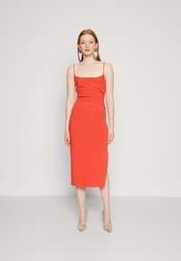 Red midi dress with thin straps, fitted silhouette, and a side slit. Worn with clear heels. Simple design, smooth texture.