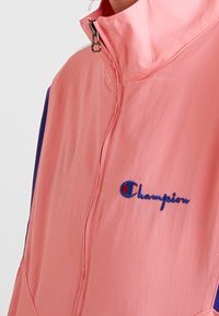 Chaqueta de cremallera rosa claro con cuello alto, con acentos en azul marino y el logo de "Champion" bordado en azul en la parte frontal. Textura suave.