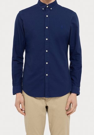 Mężczyzna ubrany w granatową koszulę na guziki z długimi rękawami i beżowe spodnie, stoi z rękami swobodnie opuszczonymi wzdłuż ciała.