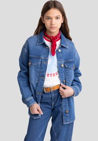 Denim jakk, millel on kaks rindtaskut, nööbid ja üleliigne sobivus. Kandes valge graafilise T-särgi kohal ja kombineerituna siniste teksadega. Punane bandaan annab värvilaiku.