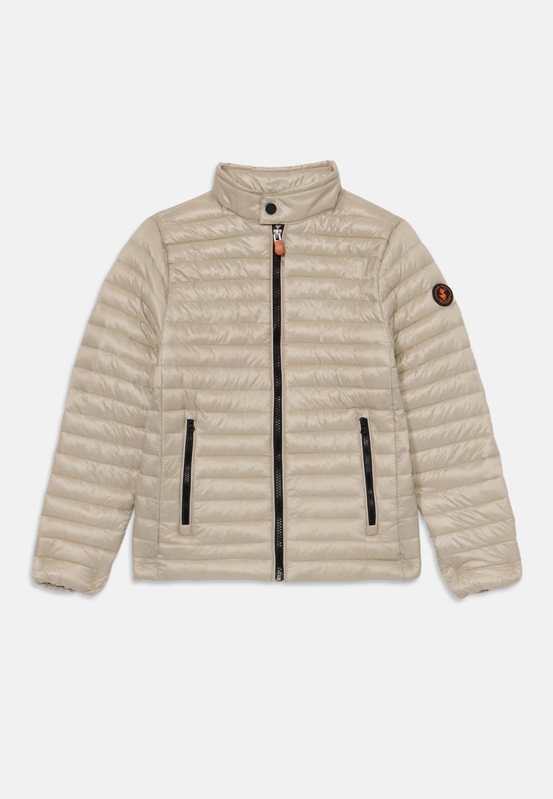 Veste matelassée beige clair avec quilting horizontal, col montant, deux poches latérales zippées noires et un patch logo sur la manche gauche.