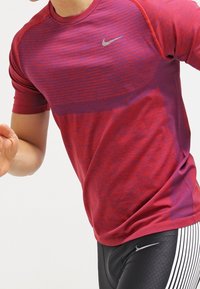 Camisa deportiva de manga corta en rojo y púrpura con un patrón a rayas, que presenta un pequeño logo de Nike en gris en el pecho. El tejido parece ligero.
