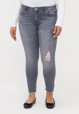 Jean skinny en denim gris délavé, avec une déchirure au genou droit, bords effilochés et design à cinq poches. Associé avec des ballerines noires.