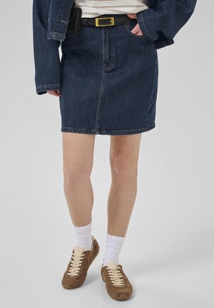 Jupe en denim noir avec une coupe droite, dotée de poches avant, associée à des baskets brunes et des chaussettes blanches. La tenue comprend une ceinture noire.