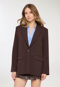 Blazer marrone dal design strutturato, con chiusura a un bottone e due tasche frontali, indossato sopra una camicia azzurro chiaro. Tessuto liscio.