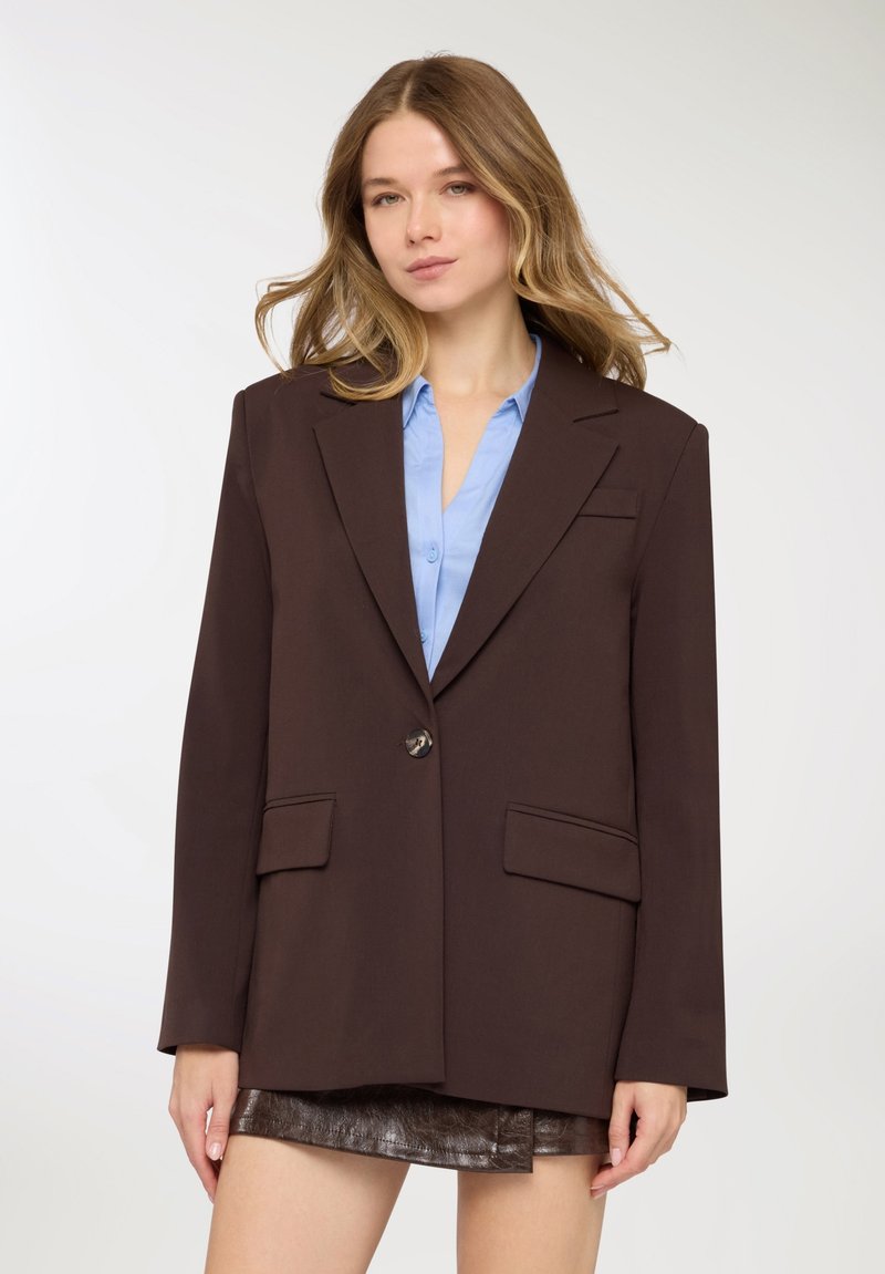 Blazer marrone dal design strutturato, con chiusura a un bottone e due tasche frontali, indossato sopra una camicia azzurro chiaro. Tessuto liscio.