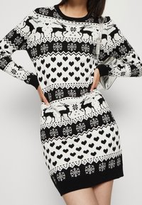 La robe en tricot présente un schéma de couleurs noir et blanc avec des motifs de rennes, de cœurs et de flocons de neige. Col rond classique et silhouette ajustée.